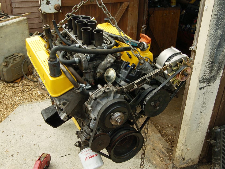 A complete V8 engine with yellow rocker covers hanging from a hoist chain outside a workshop, alternator and belts still fitted