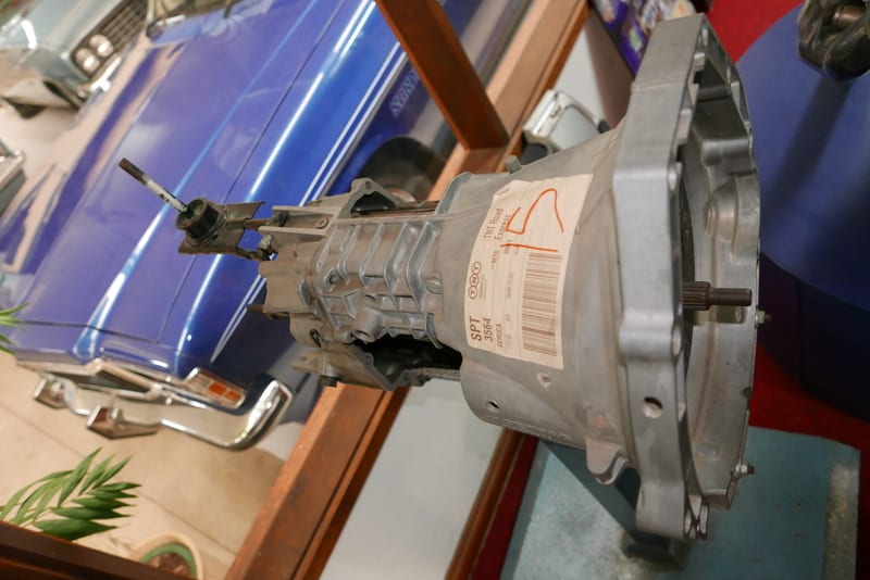 A complete five-speed manual gearbox removed from a car, resting on a bench with its bellhousing and selector visible